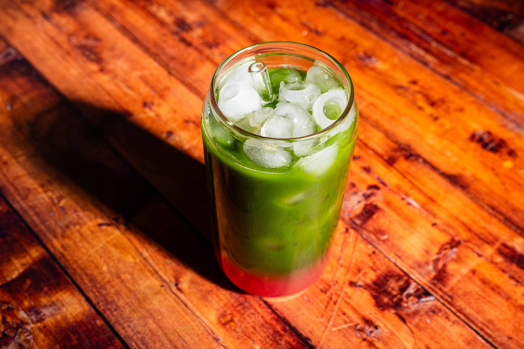 A tall glass filled with a layered green and red iced drink, topped with ice cubes, placed on a wooden surface.