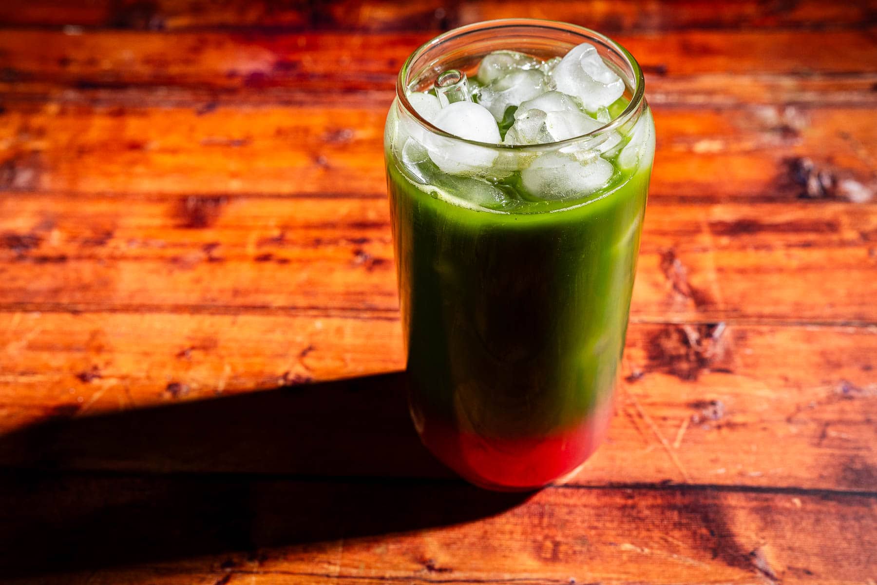 A glass filled with iced green and red layered juice sits on a wooden surface, casting a shadow to the left.
