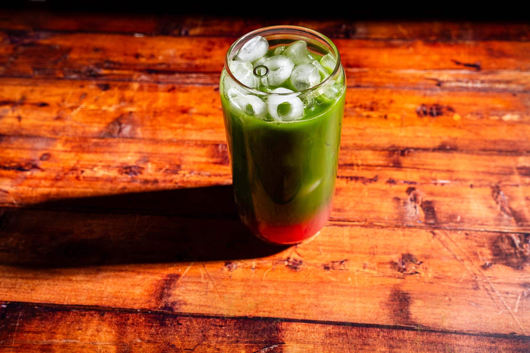 A glass of iced green and red layered drink sits on a wooden table.