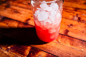 A glass filled with ice and a red-pink beverage sits on a wooden table in bright sunlight.