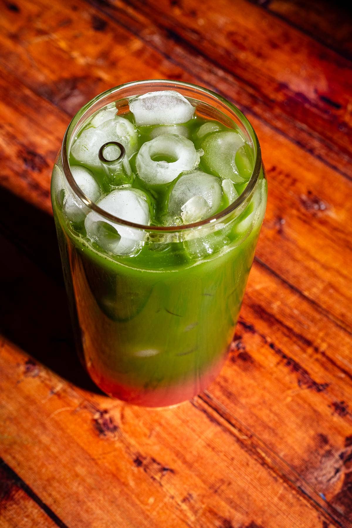 A glass filled with green juice and ice cubes sits on a wooden surface.