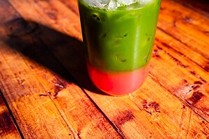 A glass of iced drink with a green top layer and a red bottom layer sits on a wooden surface in bright sunlight.