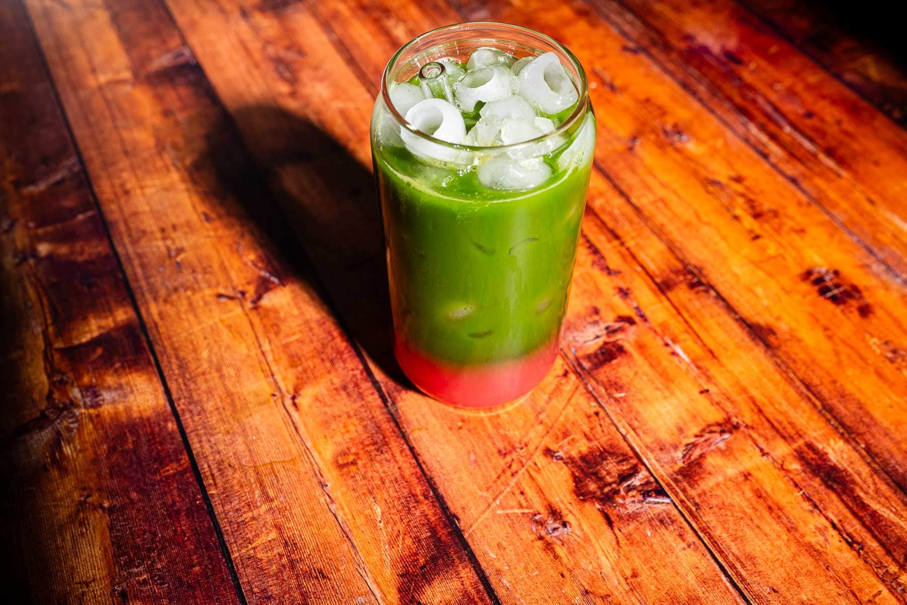 A glass of iced drink with a green liquid layered above a red liquid, set on a wooden table.