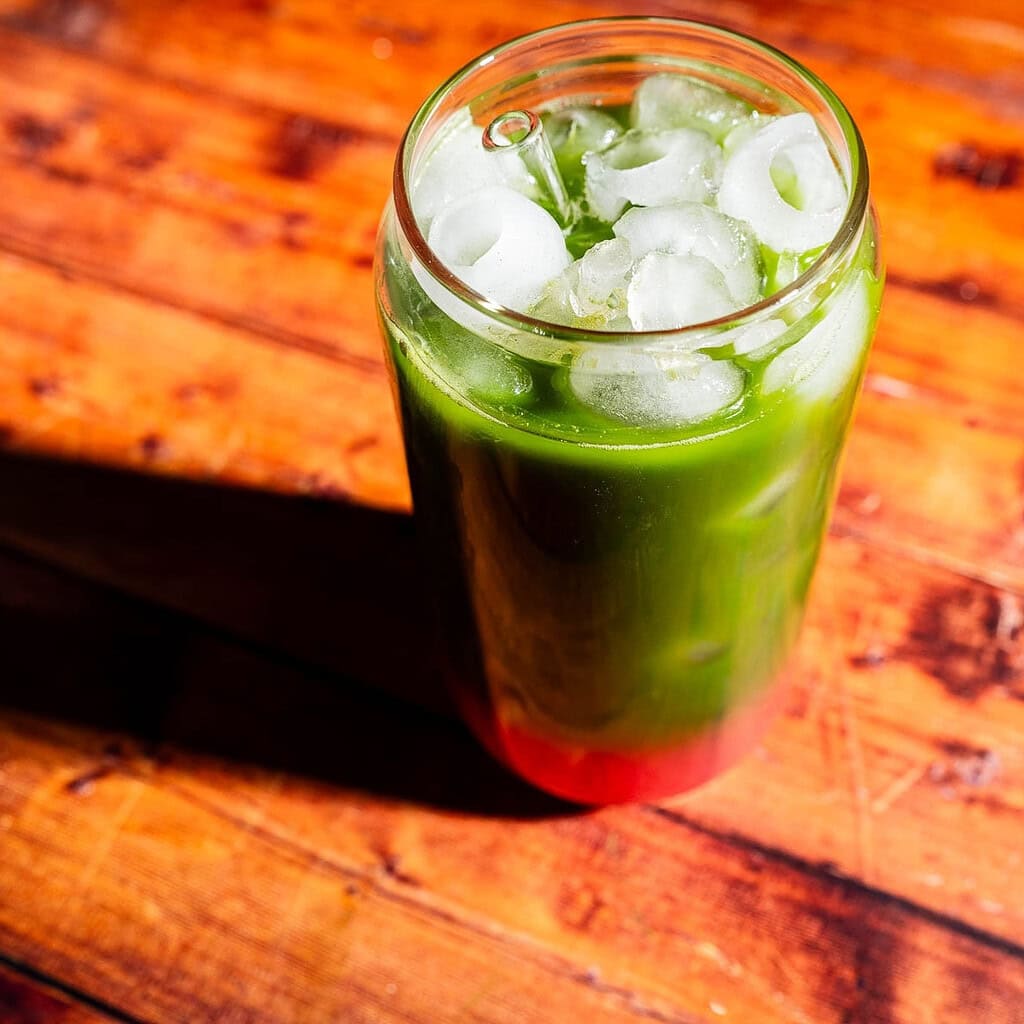 A glass of iced green beverage with red liquid at the bottom, sitting on a wooden surface in sunlight.