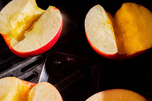 Halved apples with bright yellow flesh are placed on a dark metal baking tray, illuminated by direct light.