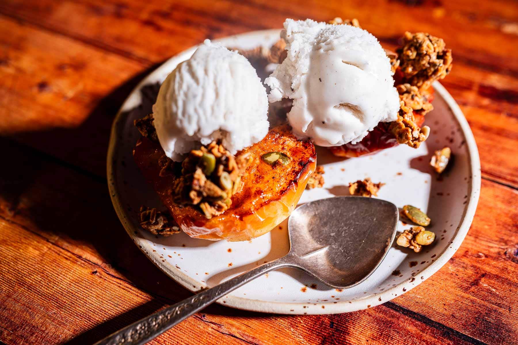 A dessert plate with two scoops of vanilla ice cream on top of air fryer baked apples and granola, next to a spoon on a wooden table.