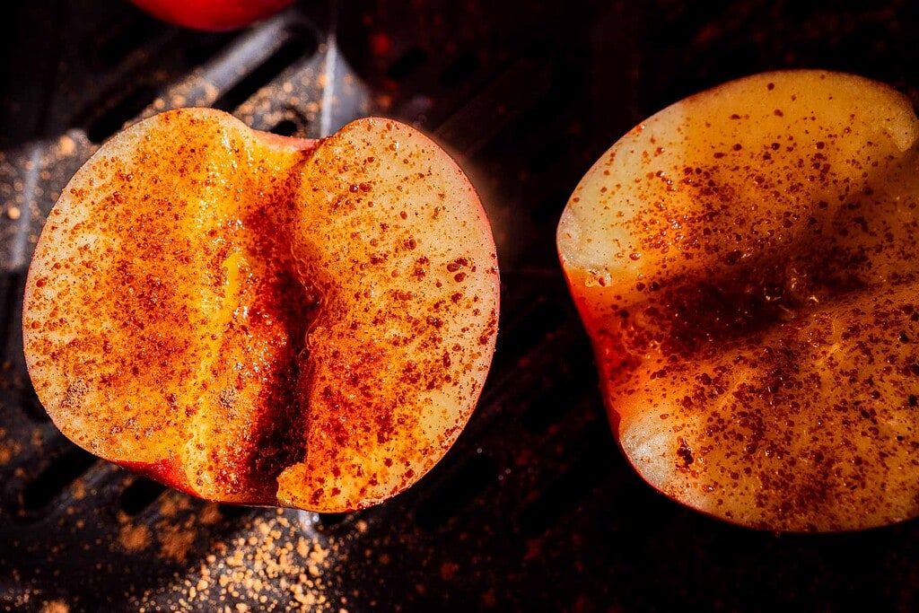 Two apple halves sprinkled with chili powder rest on a dark surface, with light highlighting the texture and spice.