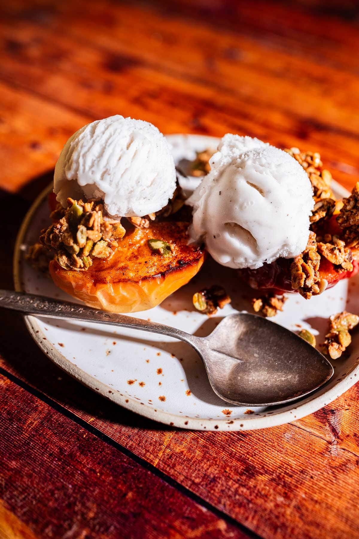 A plate with air fryer baked apples, topped with two scoops of vanilla ice cream, granola, and a spoon on the side, set on a wooden table.