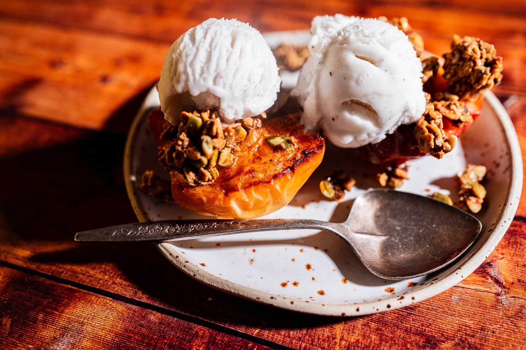 A plate with two grilled apple halves topped with scoops of vanilla ice cream and granola, reminiscent of the comforting flavors found in air fryer baked apples, sits next to a spoon on a wooden table.