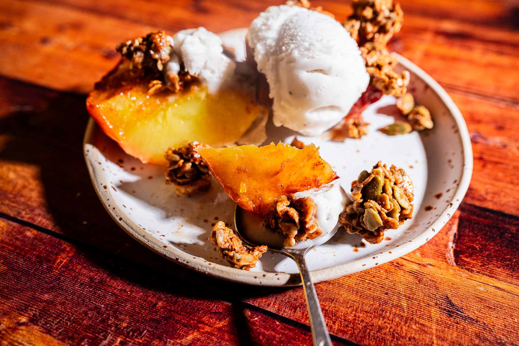A plate with baked apple halves, two scoops of vanilla ice cream, granola clusters, and a spoon, on a wooden surface.