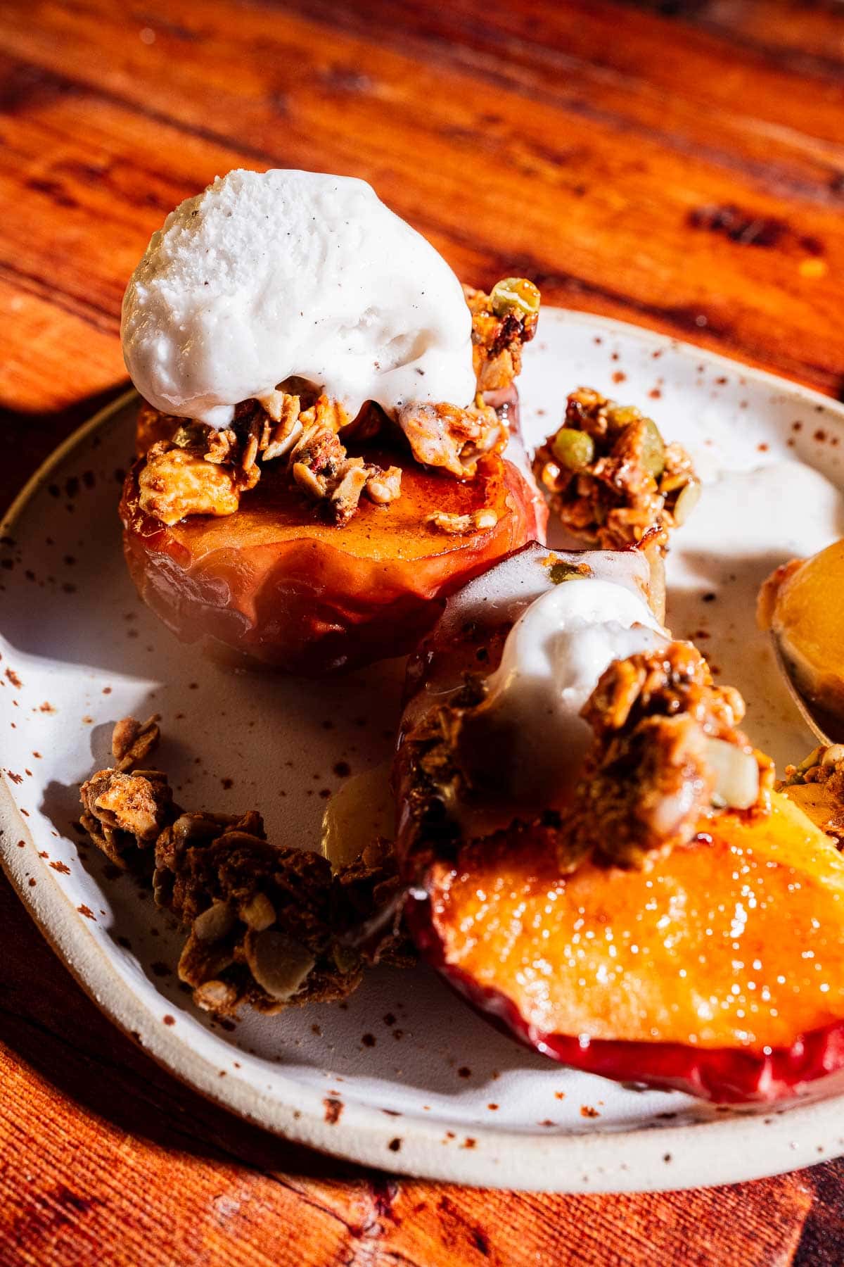 Two baked apple halves topped with granola and a scoop of vanilla ice cream on a speckled ceramic plate, set on a wooden surface.