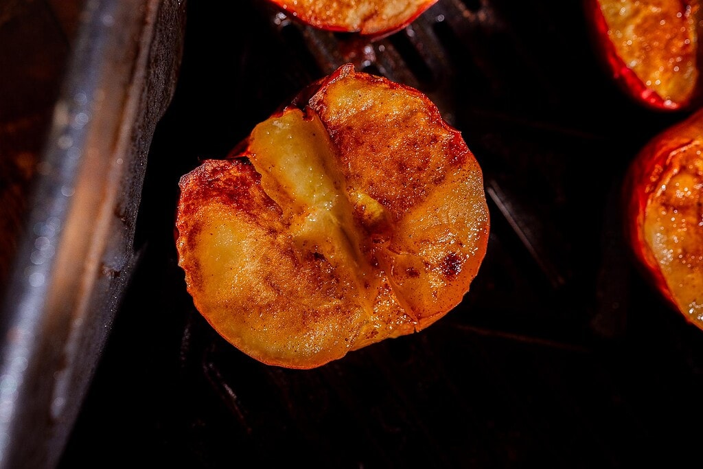 A close-up of a baked peach half with a caramelized, golden-brown surface, resting on a dark grill.