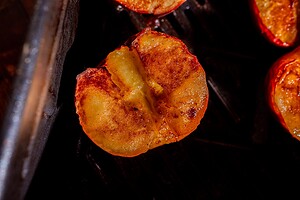 A close-up of a baked peach half with a caramelized, golden-brown surface, resting on a dark grill.