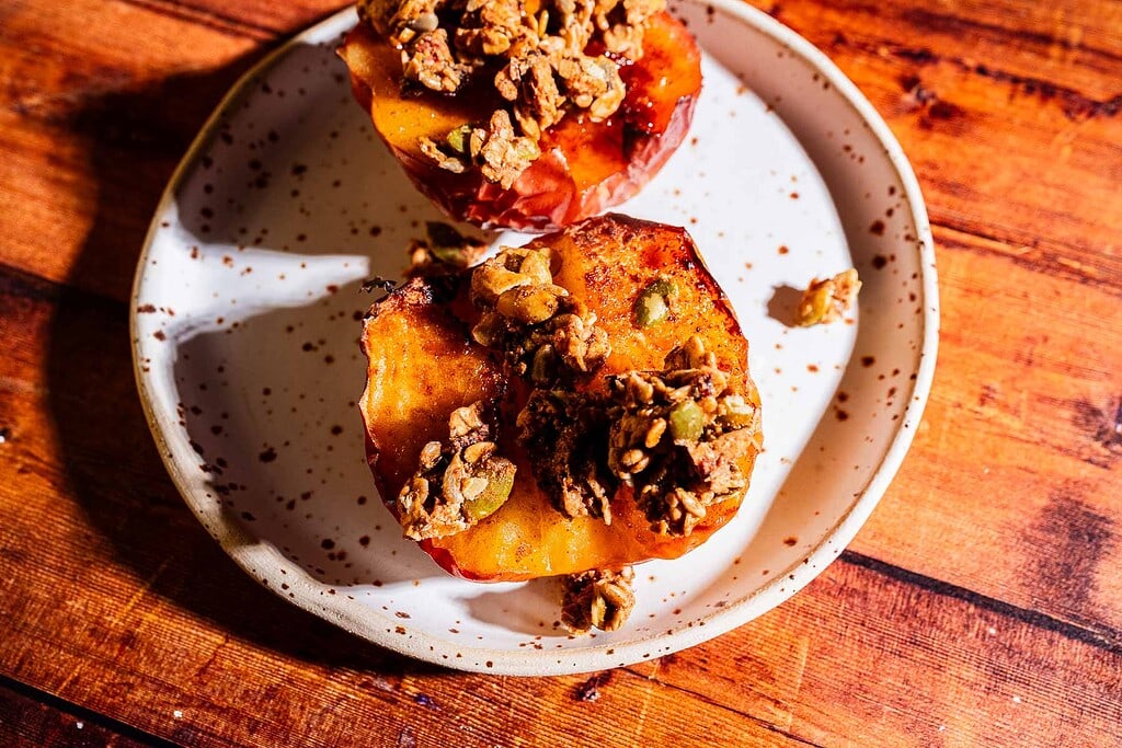 Two baked apple halves topped with granola sit on a white ceramic plate with brown speckles, placed on a wooden surface.