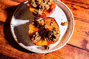 Two baked apple halves topped with granola sit on a white ceramic plate with brown speckles, placed on a wooden surface.