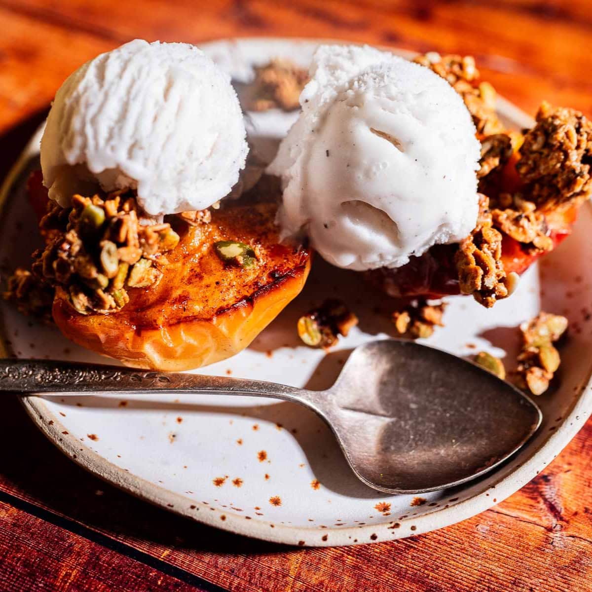 A plate with two baked apple halves topped with granola and two scoops of vanilla ice cream, next to a spoon on a rustic wooden table.