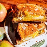 Apple cheddar grilled cheese sandwich with visible apple slices, cut in half and stacked on a plate, next to whole and sliced apples and sage leaves.