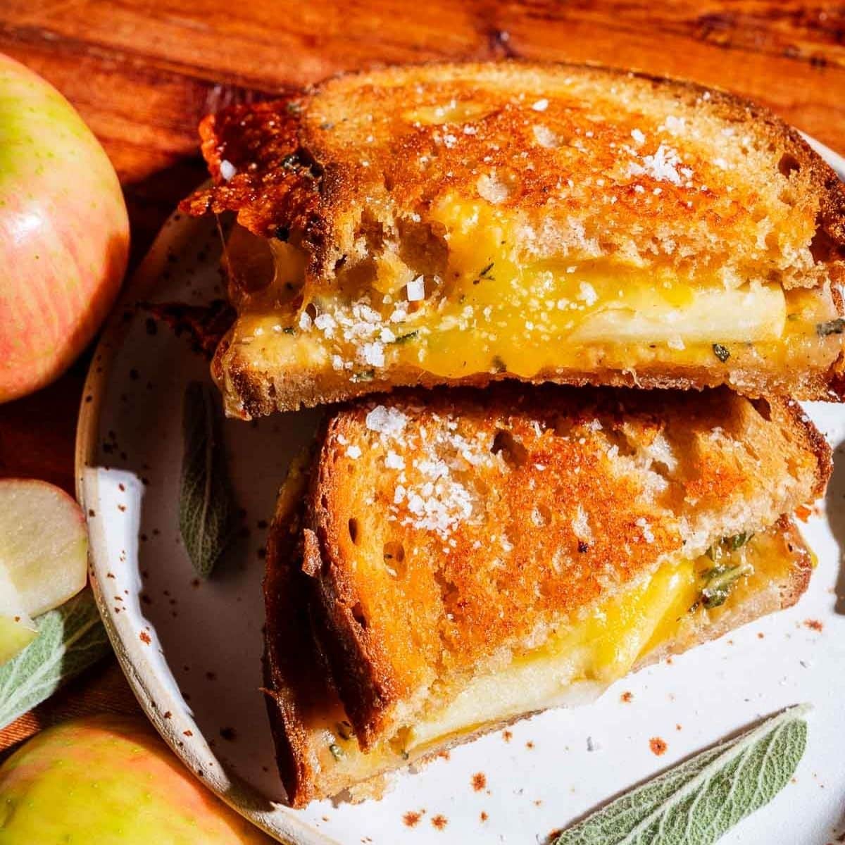 Apple cheddar grilled cheese sandwich with visible apple slices, cut in half and stacked on a plate, next to whole and sliced apples and sage leaves.