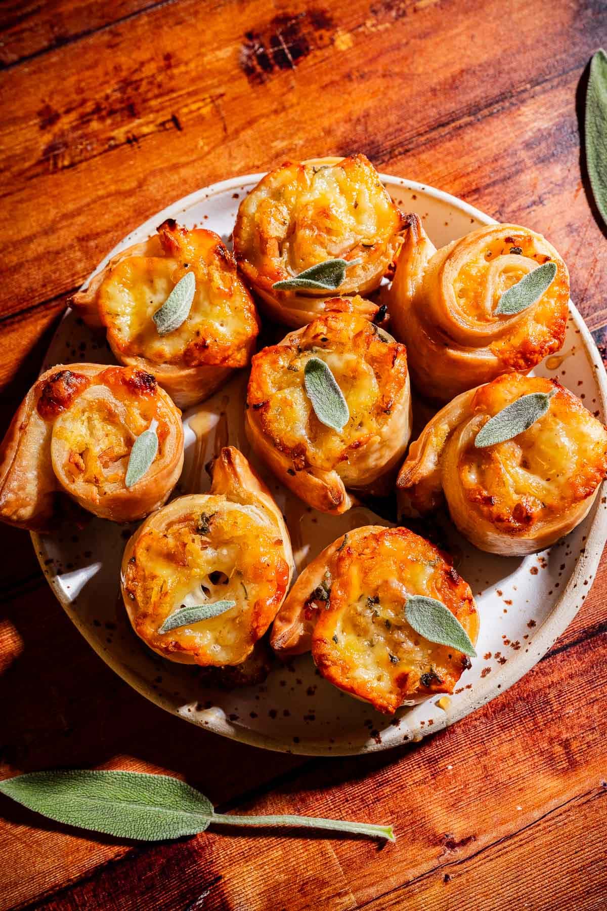 A plate of eight golden-brown savory puff pastry pinwheels topped with small sage leaves, displayed on a wooden table.
