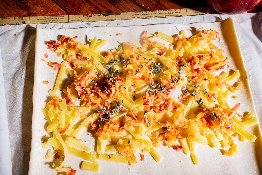 A sheet of pastry dough topped with grated apples, sliced cheese, herbs, and spices is arranged on parchment paper, ready to be rolled up into delicious puff pastry pinwheels and baked to golden perfection.