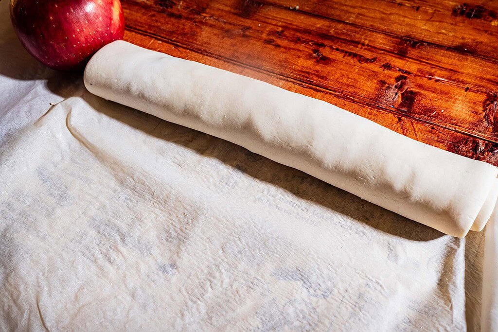 A sheet of dough is being rolled on a wooden surface next to a red apple, preparing the base for delicious puff pastry pinwheels.