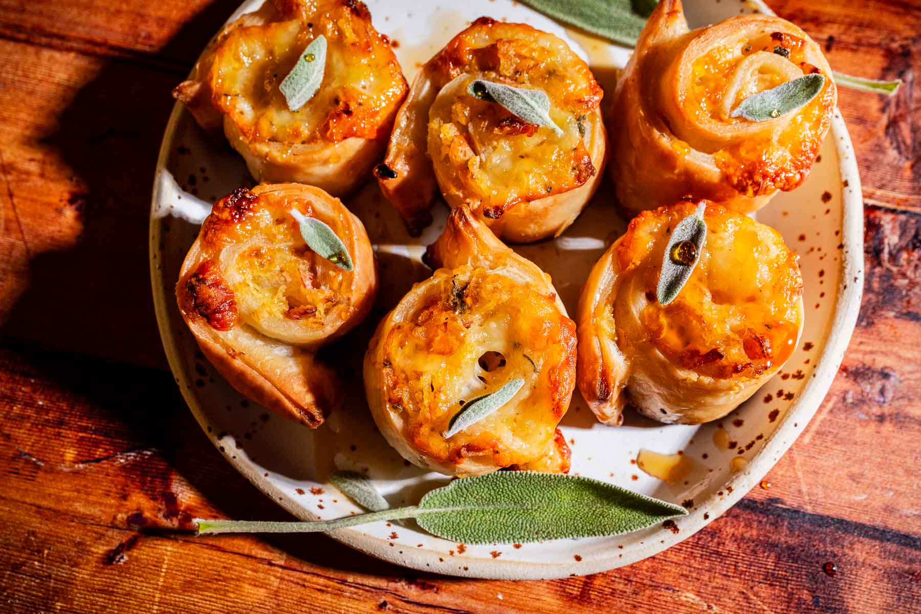 Six golden, spiral-shaped puff pastry pinwheels topped with fresh sage leaves are arranged on a ceramic plate, placed on a wooden table.