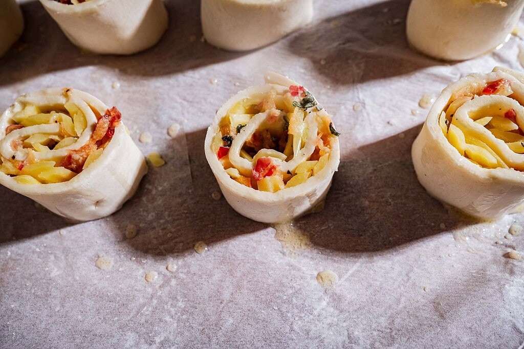 Unbaked puff pastry pinwheels with cheese, herbs, and tomato filling are arranged on a floured surface, ready for baking.