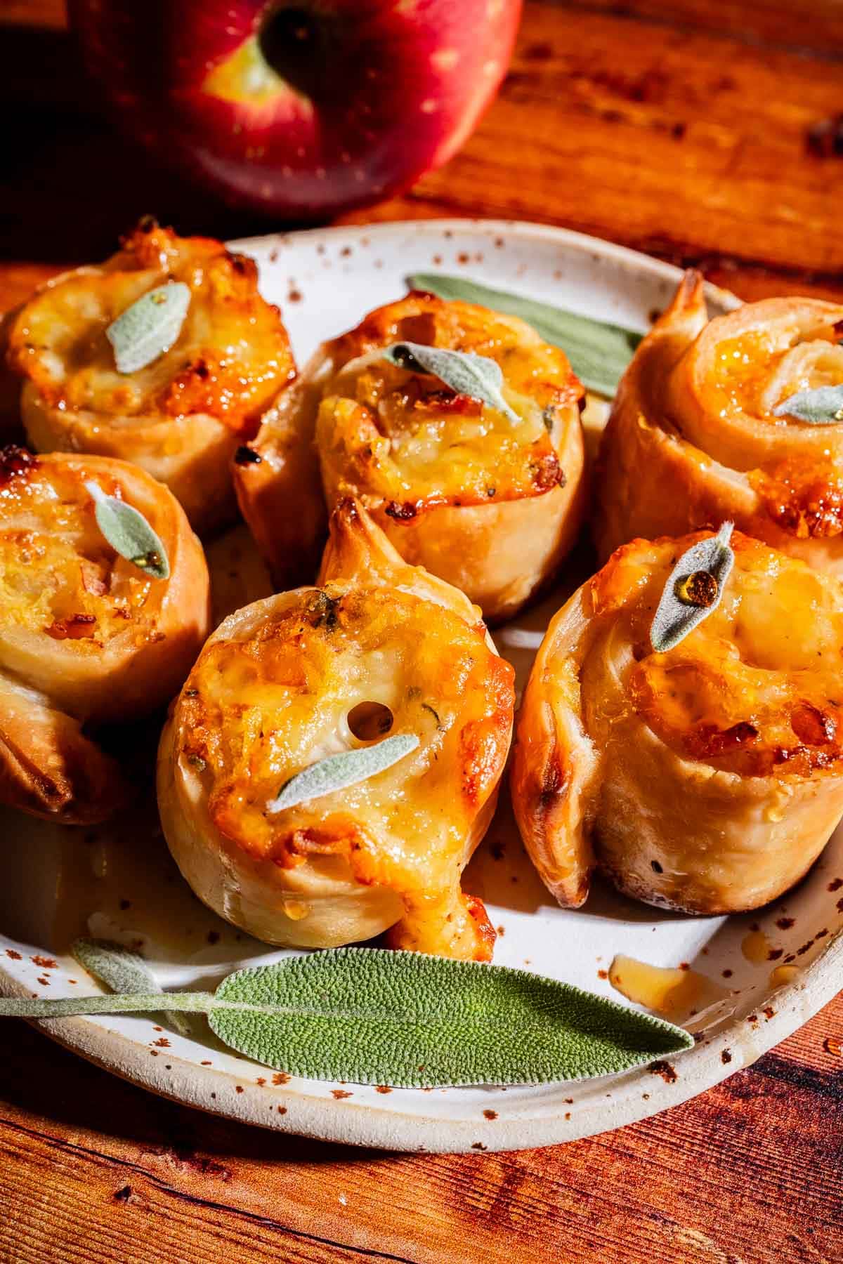 A plate of five golden puff pastry pinwheels with melted cheese and herbs, garnished with fresh sage leaves, sits on a wooden table with an apple in the background.