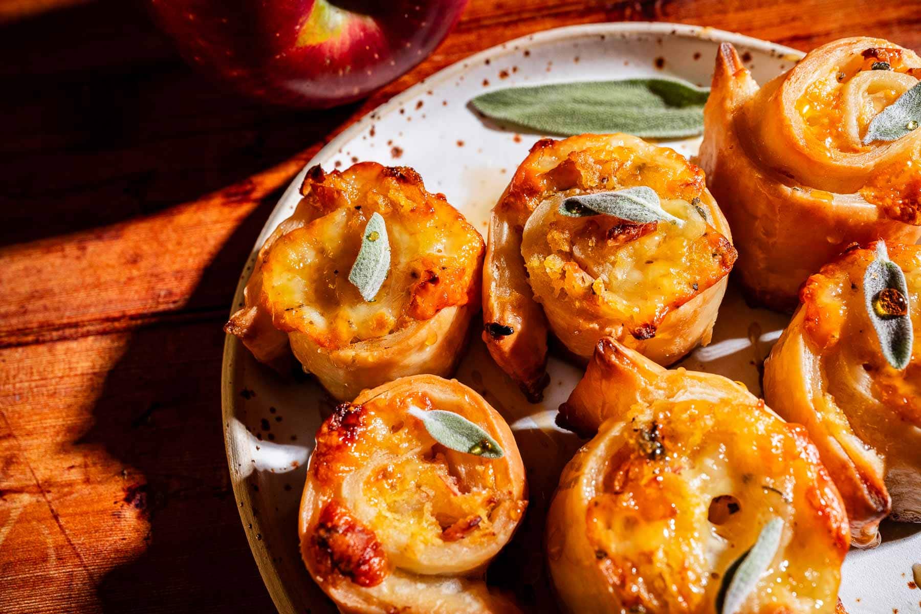 Golden puff pastry pinwheels filled with cheese and garnished with fresh sage leaves are arranged on a speckled plate next to a red apple on a wooden surface.
