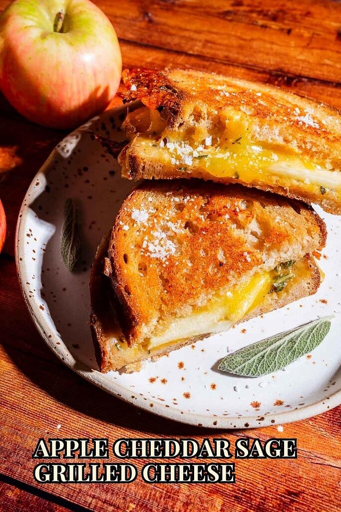 A delicious apple cheddar grilled cheese sandwich featuring sharp cheddar, crisp apple slices, and fragrant sage is cut in half on a plate, with fresh apples in the background. Text reads: “Apple Cheddar Sage Grilled Cheese.”.
