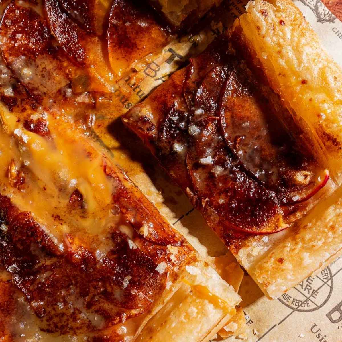 Close-up of a sliced caramel apple tart with flaky crust, caramelized apple slices, and a sprinkle of sea salt on brown parchment paper.