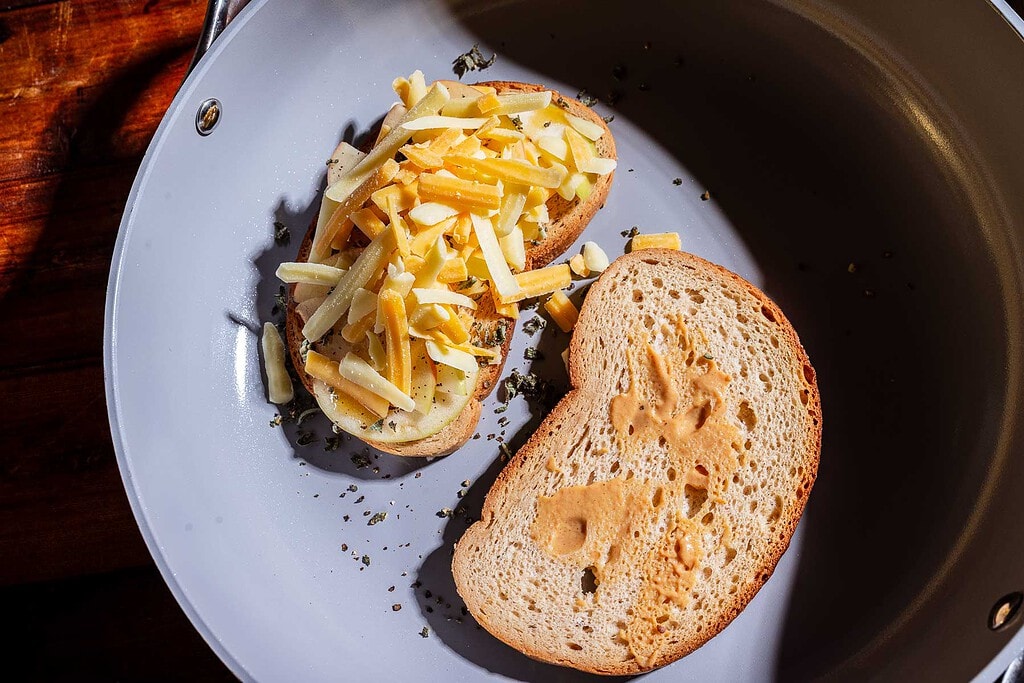 Two slices of bread in a pan, one topped with shredded cheese, ready to be cooked into a delicious apple cheddar grilled cheese.