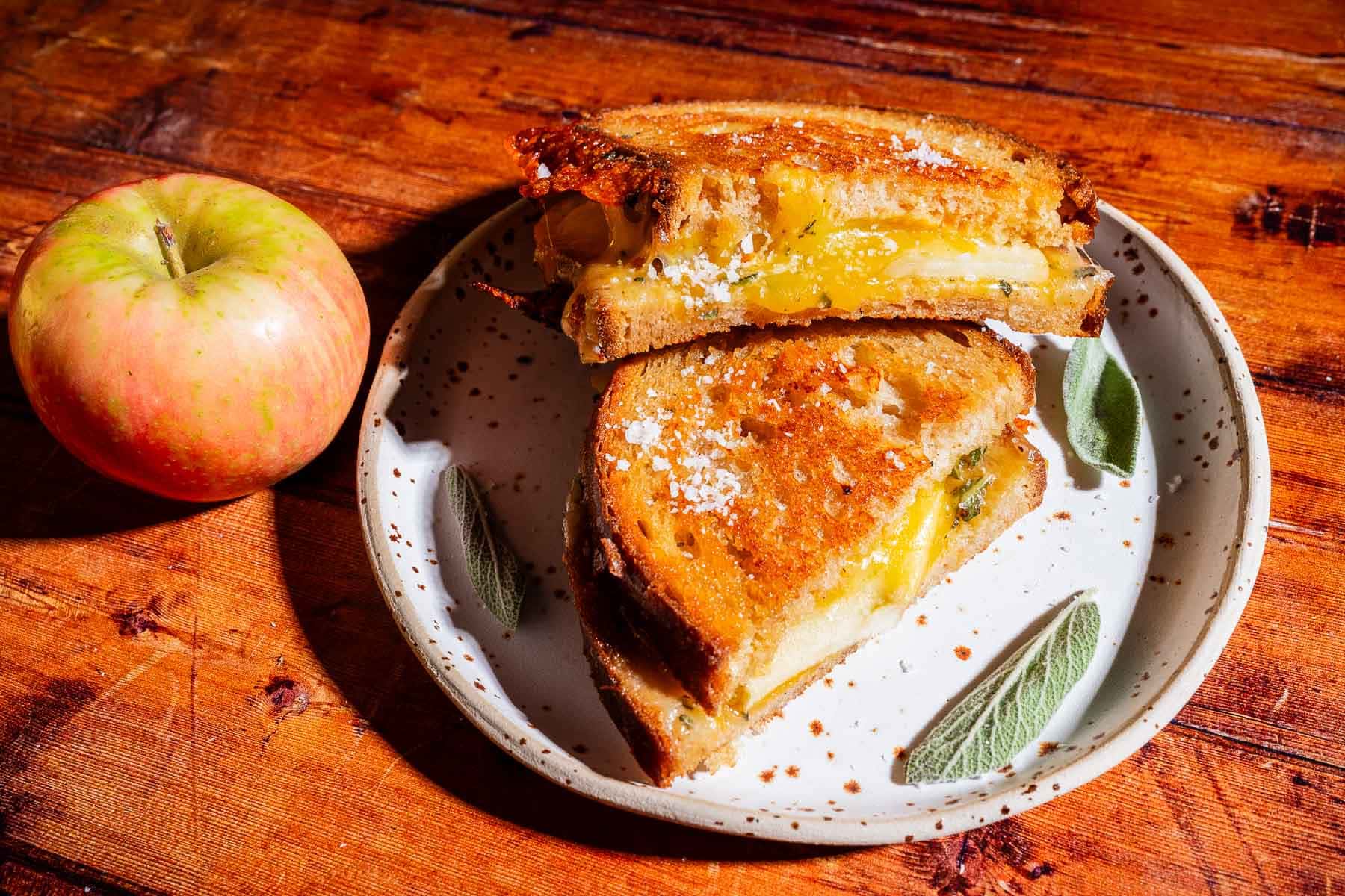 An apple cheddar grilled cheese sandwich, cut in half, sits on a speckled plate with green leaf designs, next to a whole apple on a wooden table.