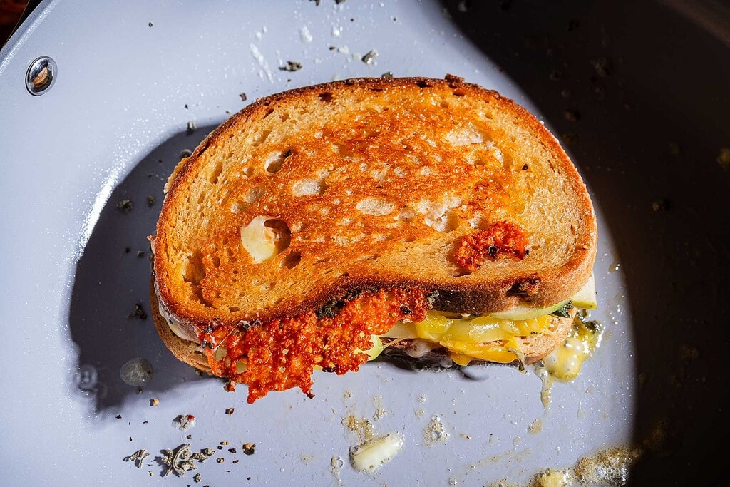 An apple cheddar grilled cheese sandwich with melted cheese spilling out sits in a white frying pan, with bits of herbs and crisp edges visible.