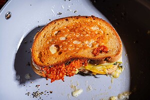 An apple cheddar grilled cheese sandwich with melted cheese spilling out sits in a white frying pan, with bits of herbs and crisp edges visible.