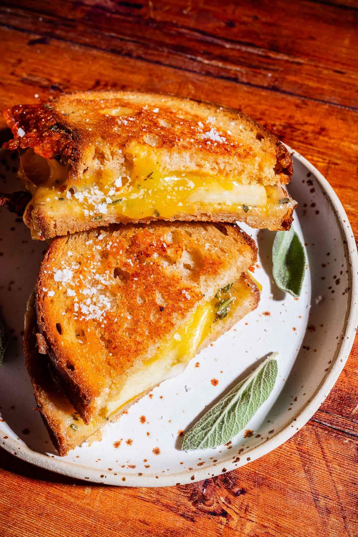 An apple cheddar grilled cheese sandwich, cut in half with melted cheese oozing out, sits on a plate with a leaf pattern on a wooden table.