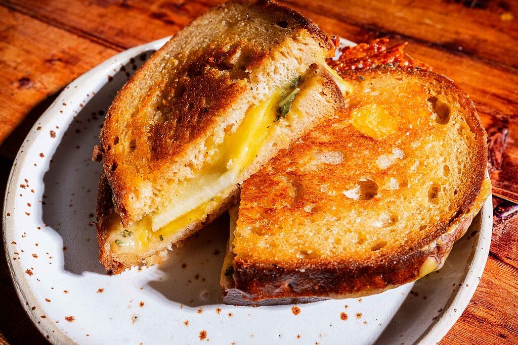 A golden apple cheddar grilled cheese sandwich cut in half on a white plate, displaying perfectly toasted bread and gooey melted cheese inside.