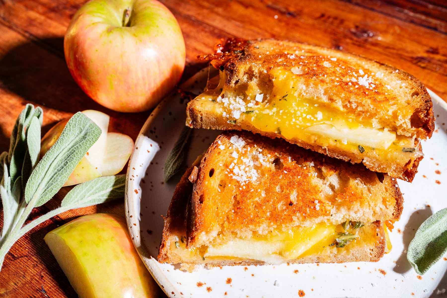An apple cheddar grilled cheese sandwich with apple slices on a plate, accompanied by an apple and sage leaves on a wooden surface nearby.