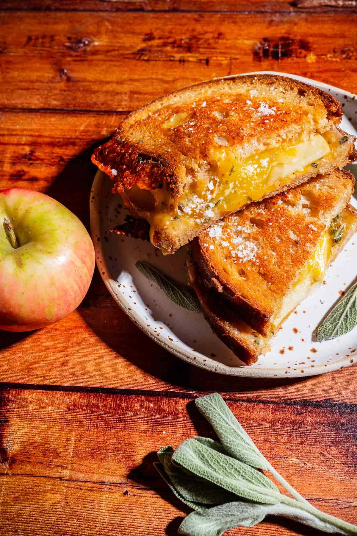 A delicious apple cheddar grilled cheese sandwich, cut in half on a plate, sits beside a whole apple and some sage leaves, all arranged on a wooden surface.