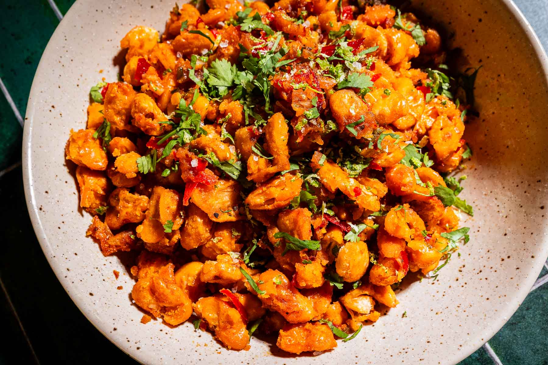A beige bowl filled with cooked, seasoned gnocchi pieces and crispy butter beans, garnished with chopped cilantro and red pepper flakes, sits on a dark green surface.