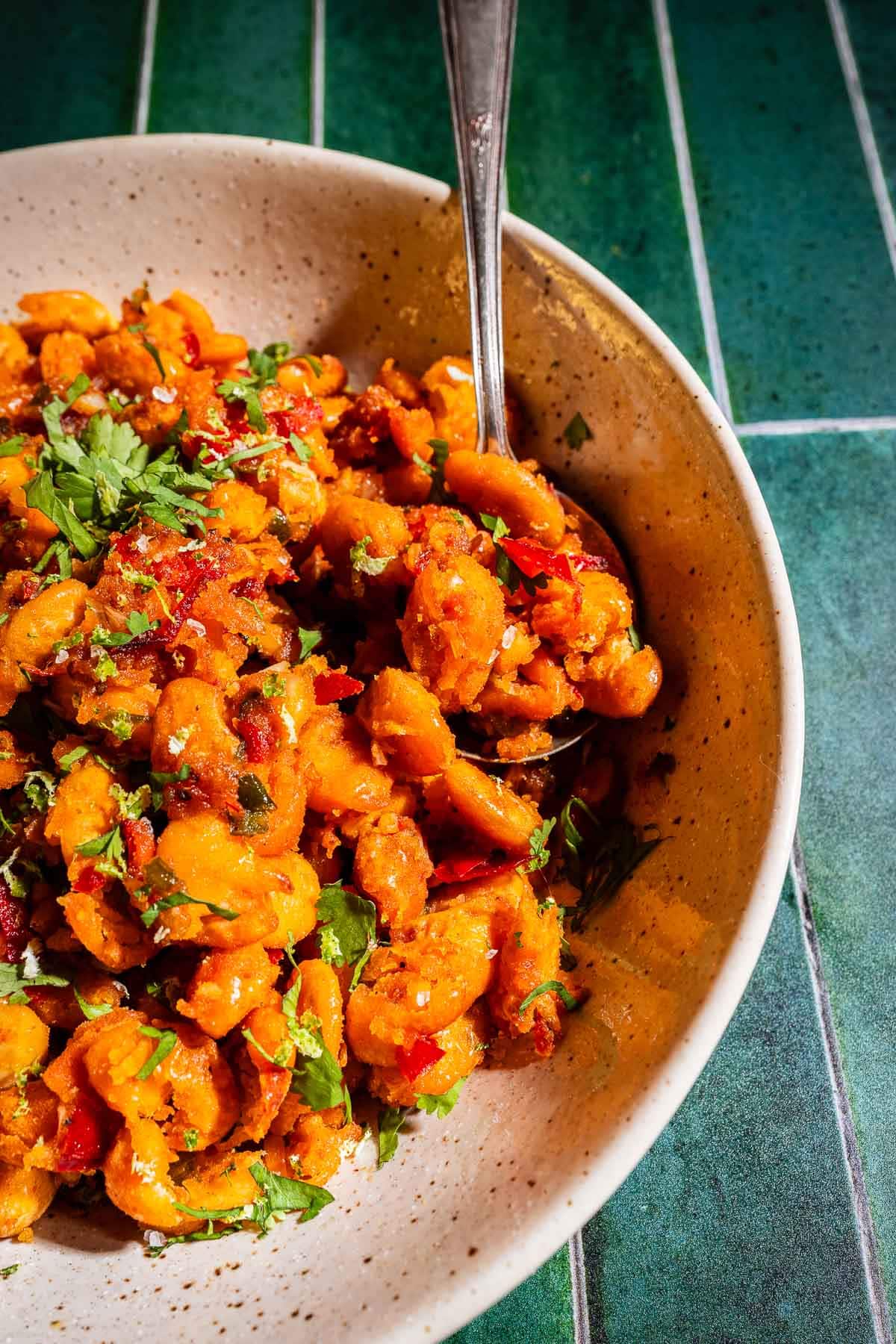 A bowl of seasoned, sautéed crispy butter beans garnished with fresh herbs, with a spoon resting in the dish, on a green tiled surface.