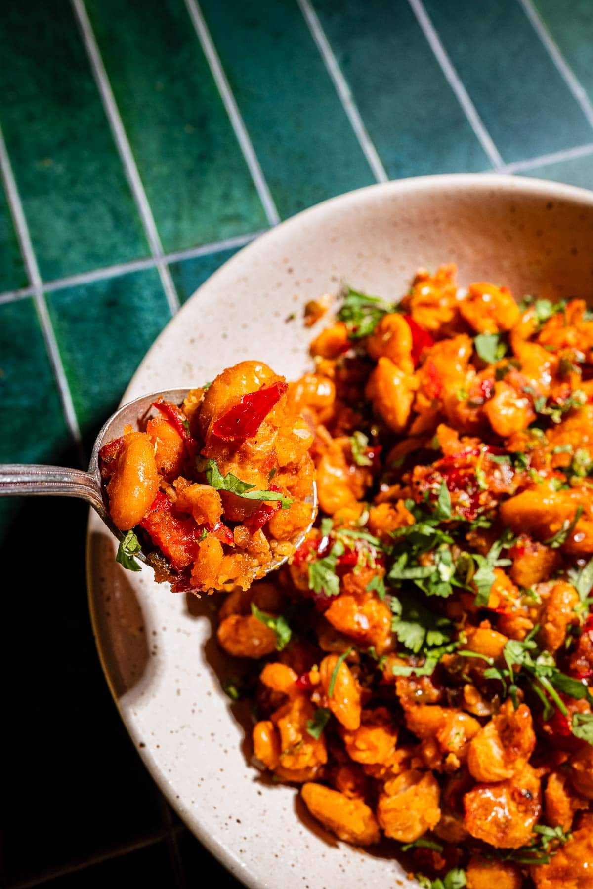 A close-up of a spoon holding spicy, crispy butter beans tossed with chopped herbs and red peppers, hovering over a bowl of the same savory dish on a green tiled surface.
