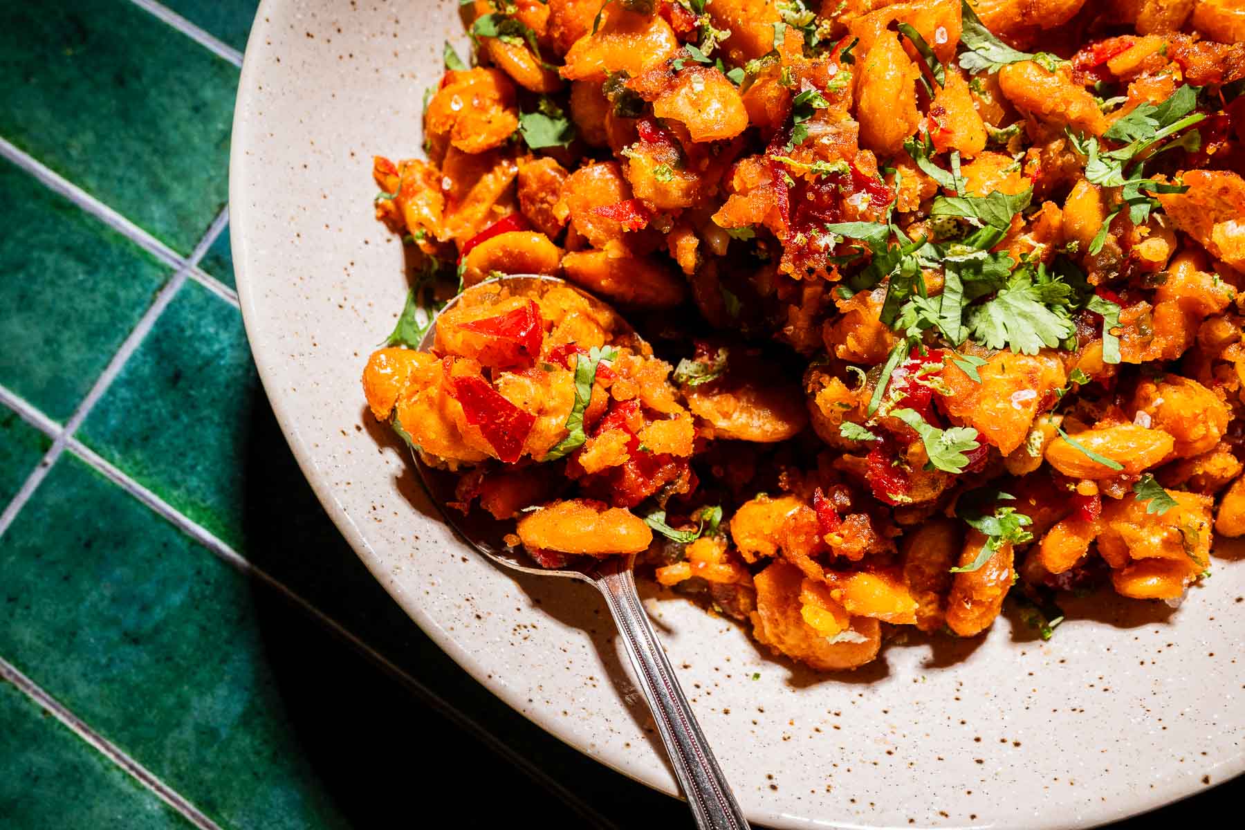A close-up of a plate with crispy butter beans mixed with herbs and red peppers, garnished with fresh cilantro, on a green tiled surface.