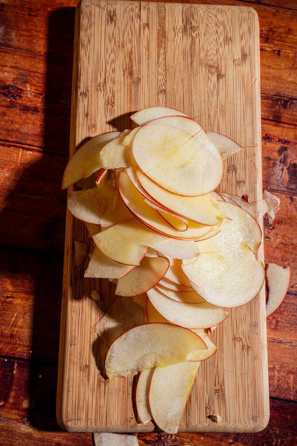Thinly sliced apple pieces are arranged on a wooden cutting board placed on a wooden surface, ready to be transformed into a delicious caramel apple tart.