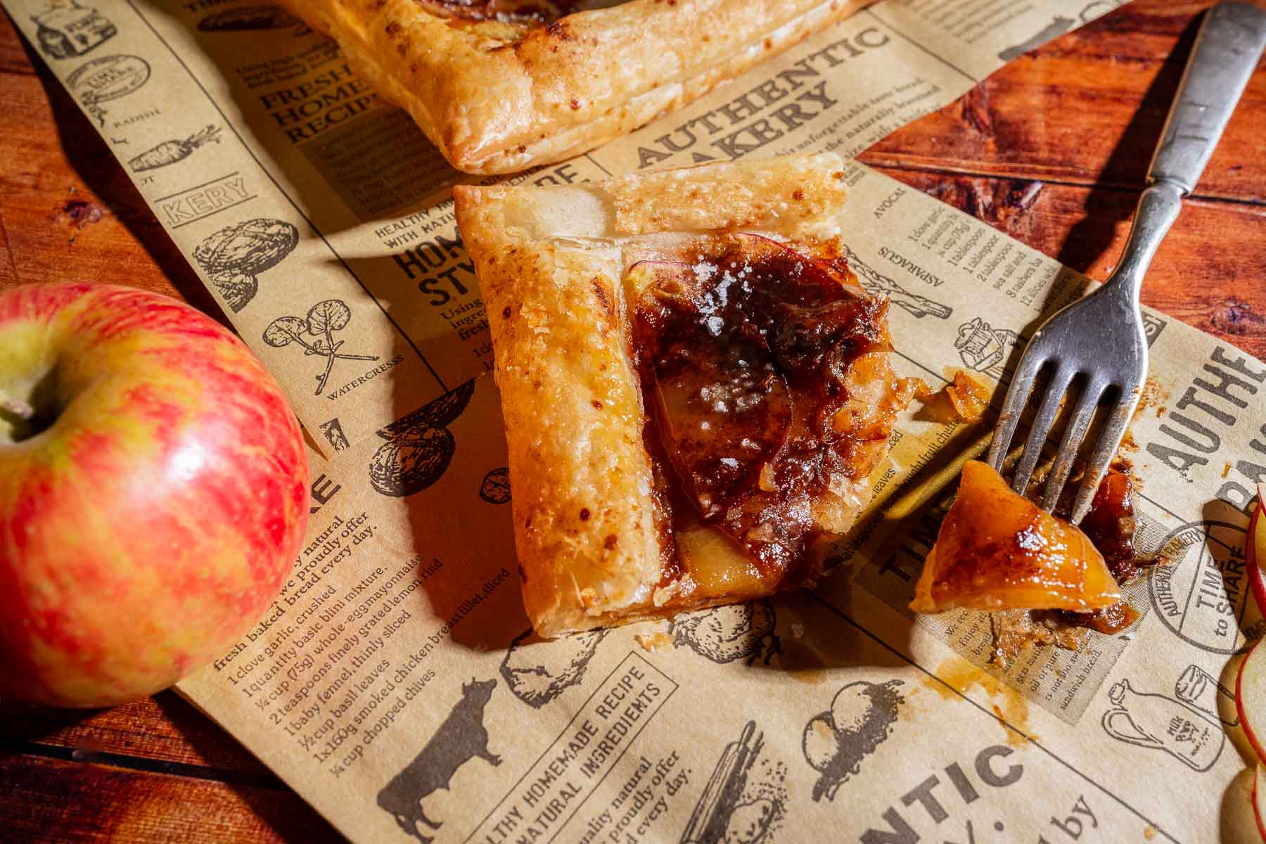 Square slice of caramel apple tart with caramelized filling on rustic printed paper beside a whole apple and a fork.