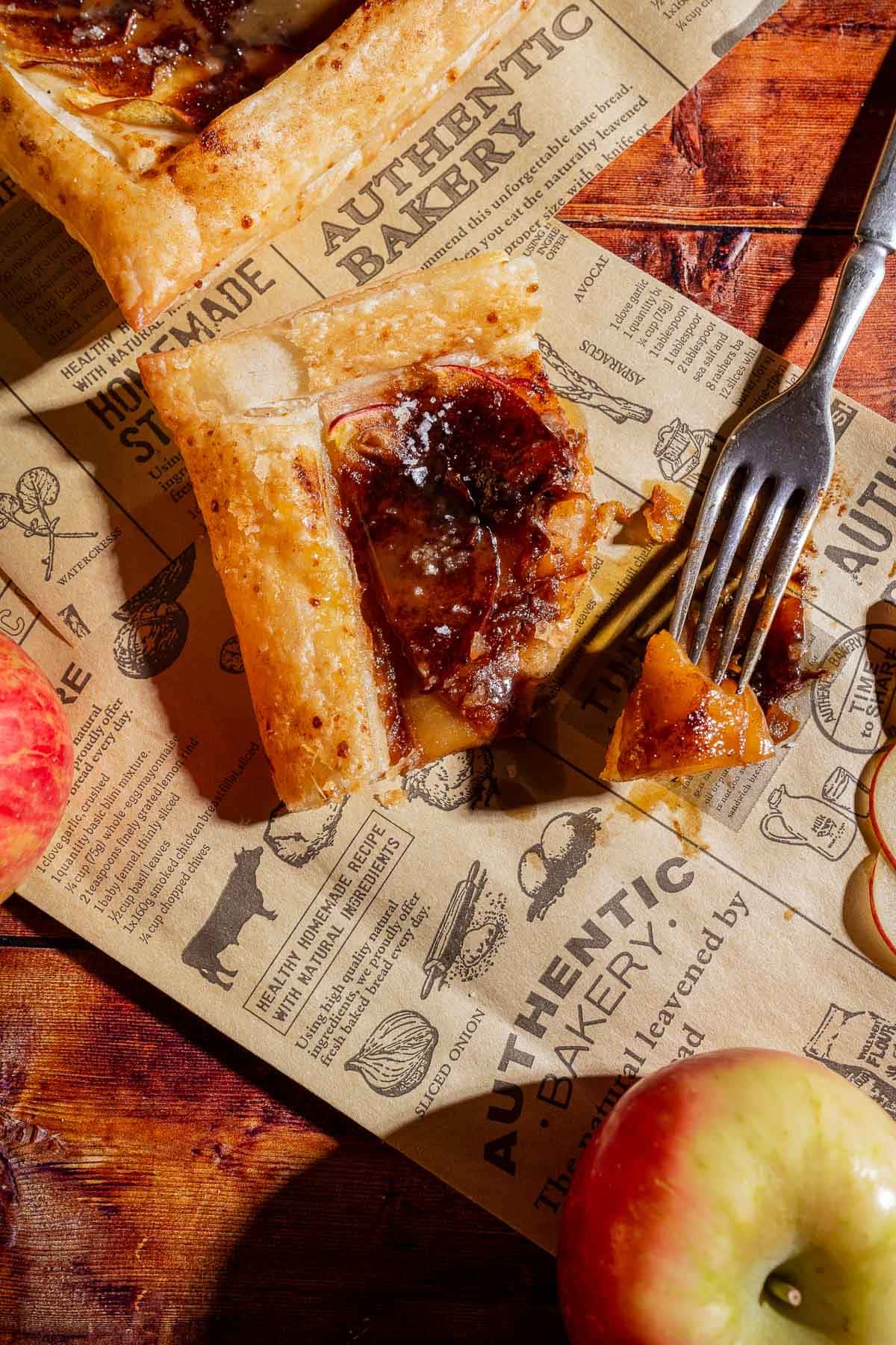 A slice of caramel apple tart sits on bakery-themed parchment paper, with a fork holding a bite-sized piece. Apples and thin apple slices are nearby on a wooden surface.