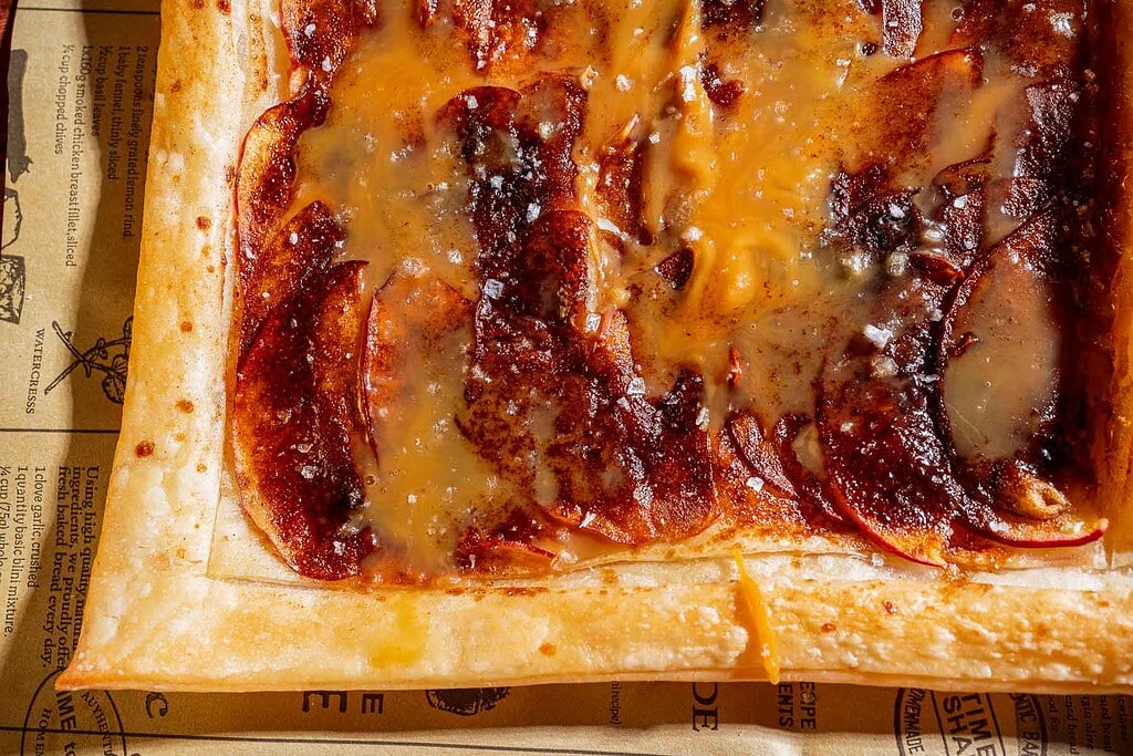 Close-up of a rectangular caramel apple tart with cinnamon, sugar, and melted cheese on a flaky pastry crust, resting on brown parchment paper.