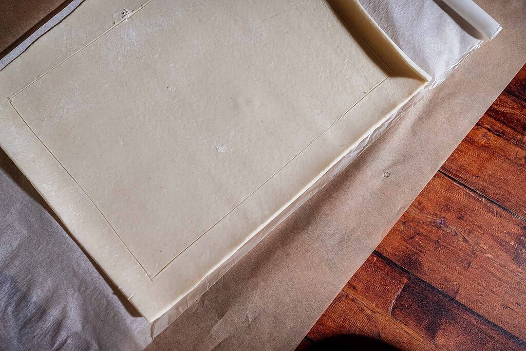 Rectangular sheet of pastry dough on parchment paper, with a scored border, placed on a wooden surface—perfectly prepared for crafting a delicious caramel apple tart.