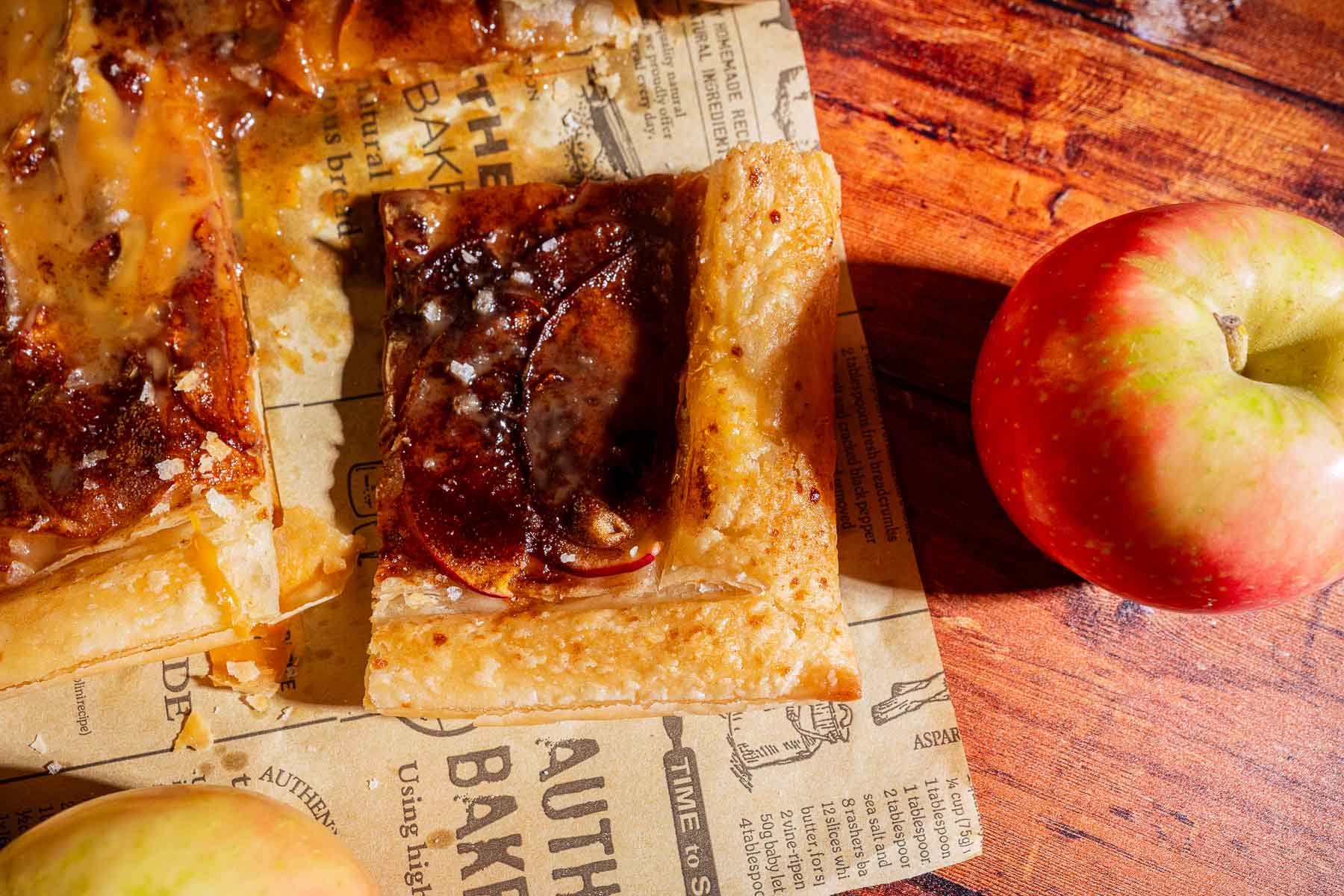 A slice of caramel apple tart with caramelized topping rests on parchment paper, next to a whole red and green apple, on a rustic wooden surface.