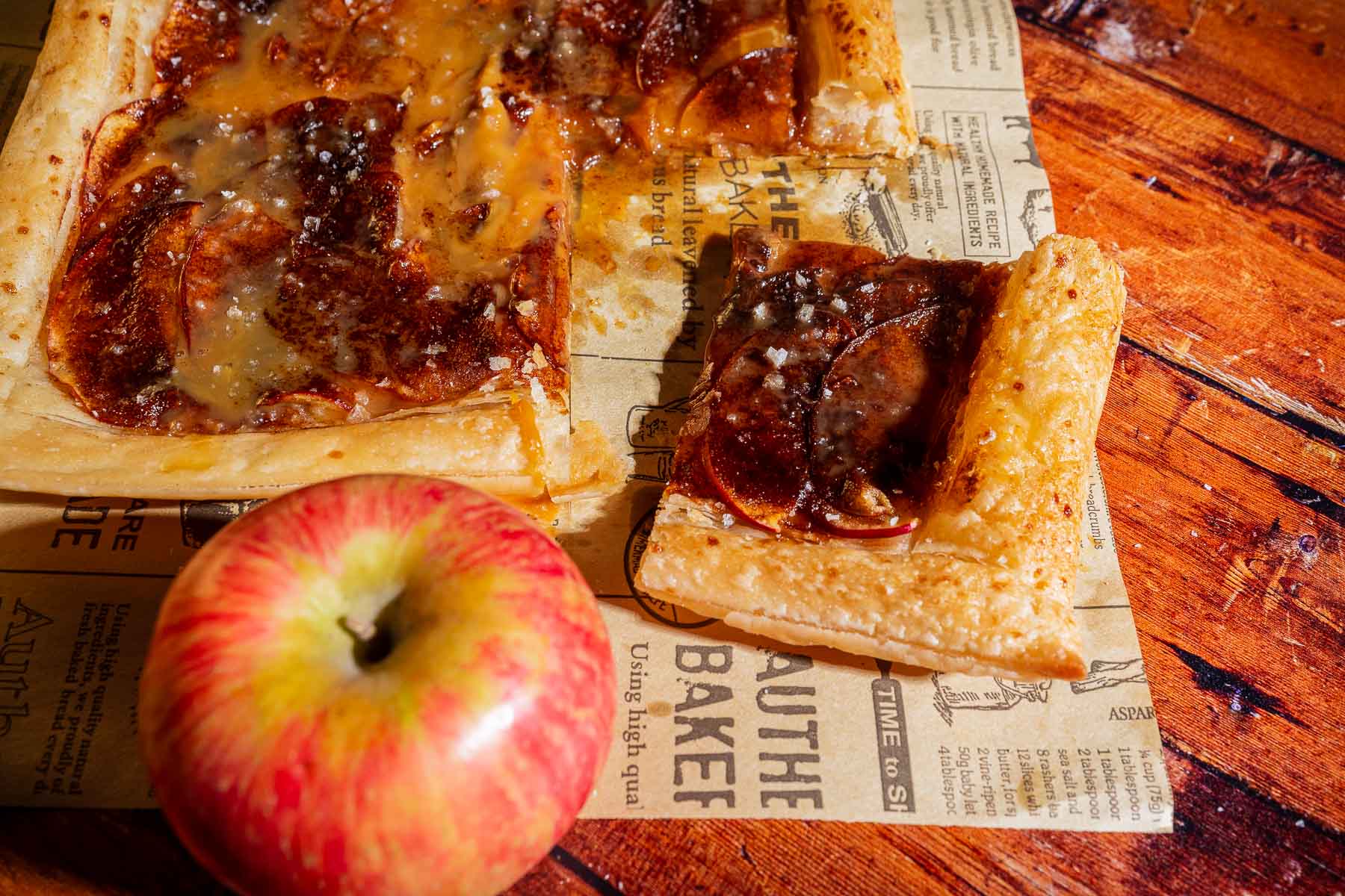 A sliced caramel apple tart with a glossy caramel glaze sits on parchment paper beside a whole red apple on a wooden surface.